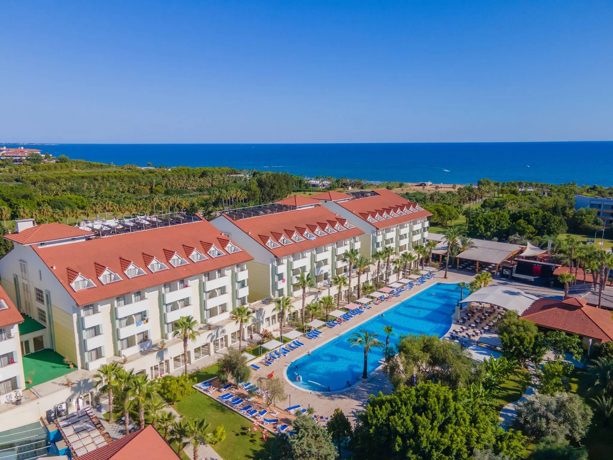 Хотелски корпуси с червени покриви и басейн, на фона на морето – Sural Resort, Сиде