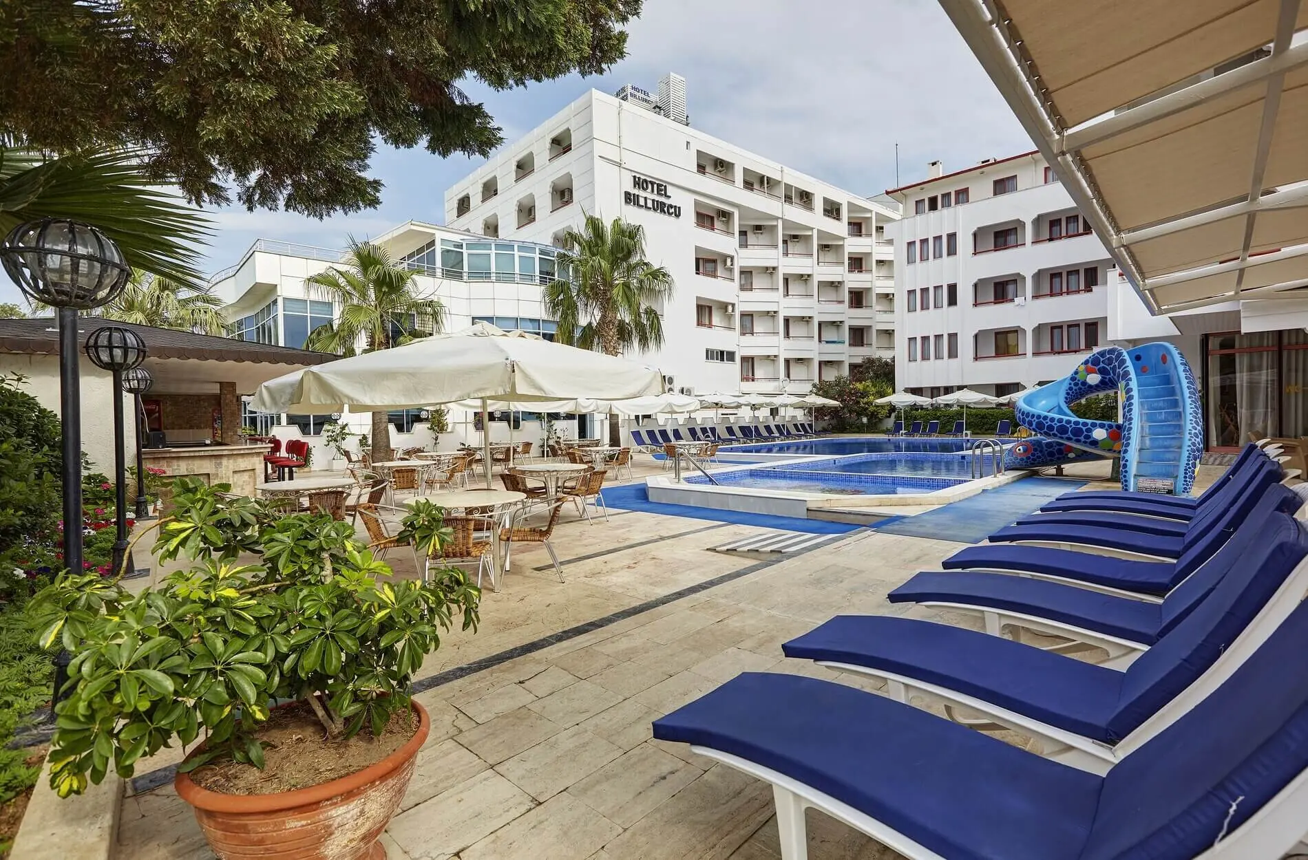 Басейн със сини шезлонги и водна пързалка пред сградата на хотела – Hotel Billurcu, Айвалък
