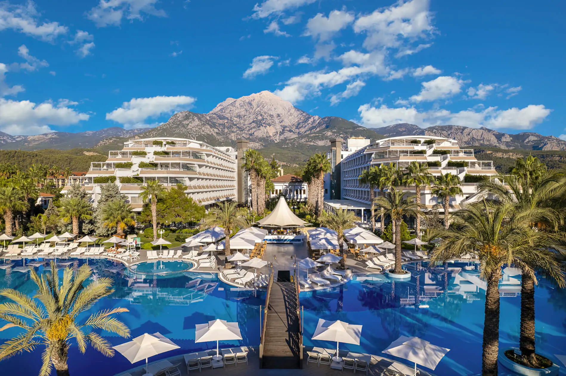 Басейн с мост, сградите и планината Тахталъ зад тях – Rai Premium Tekirova (ex.Queens Park Resort), Кемер