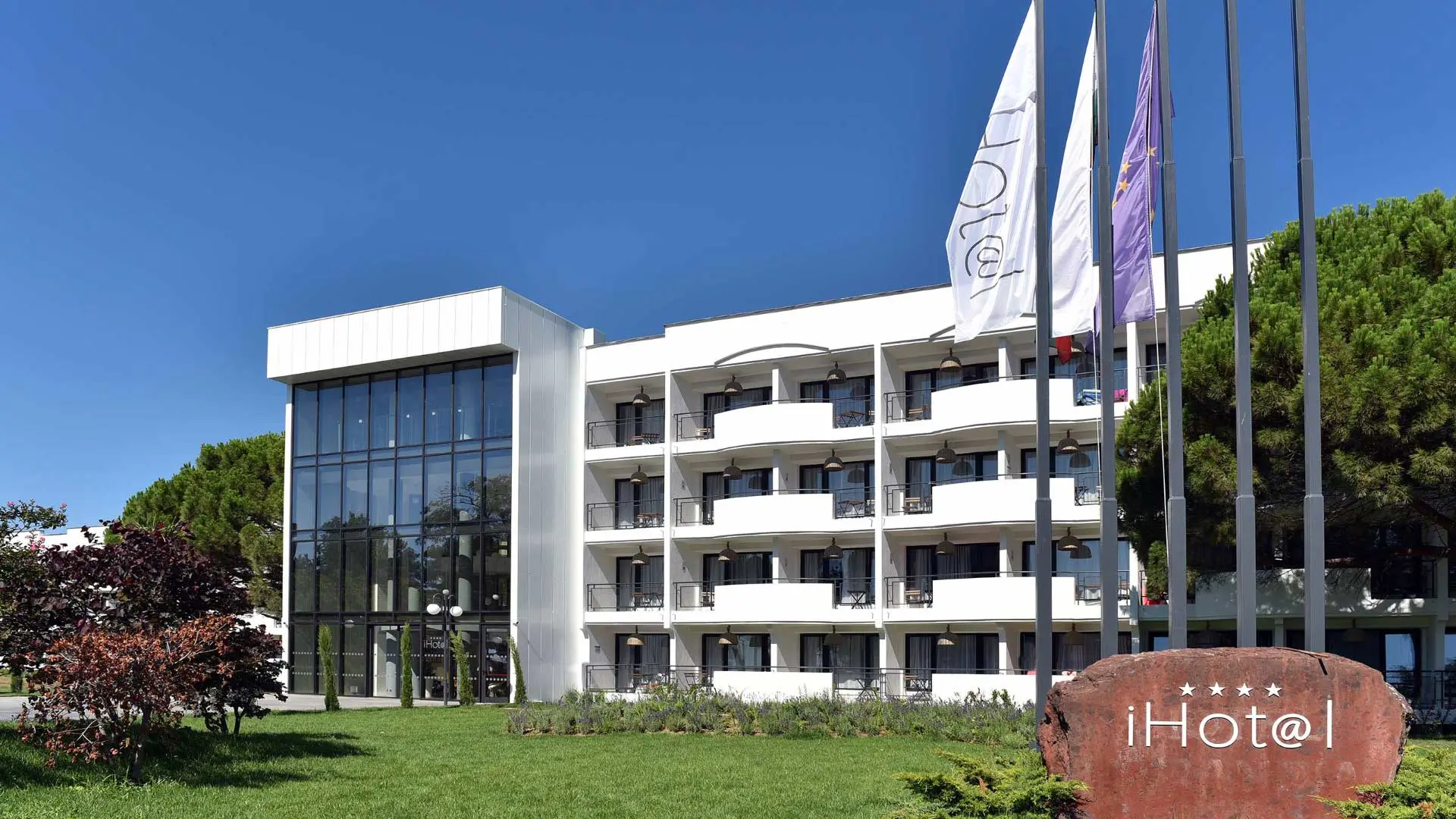 Бялата сграда на хотела със знамена, зелена морава и каменна табела с логото – iHotel Sunny Beach, Слънчев бряг