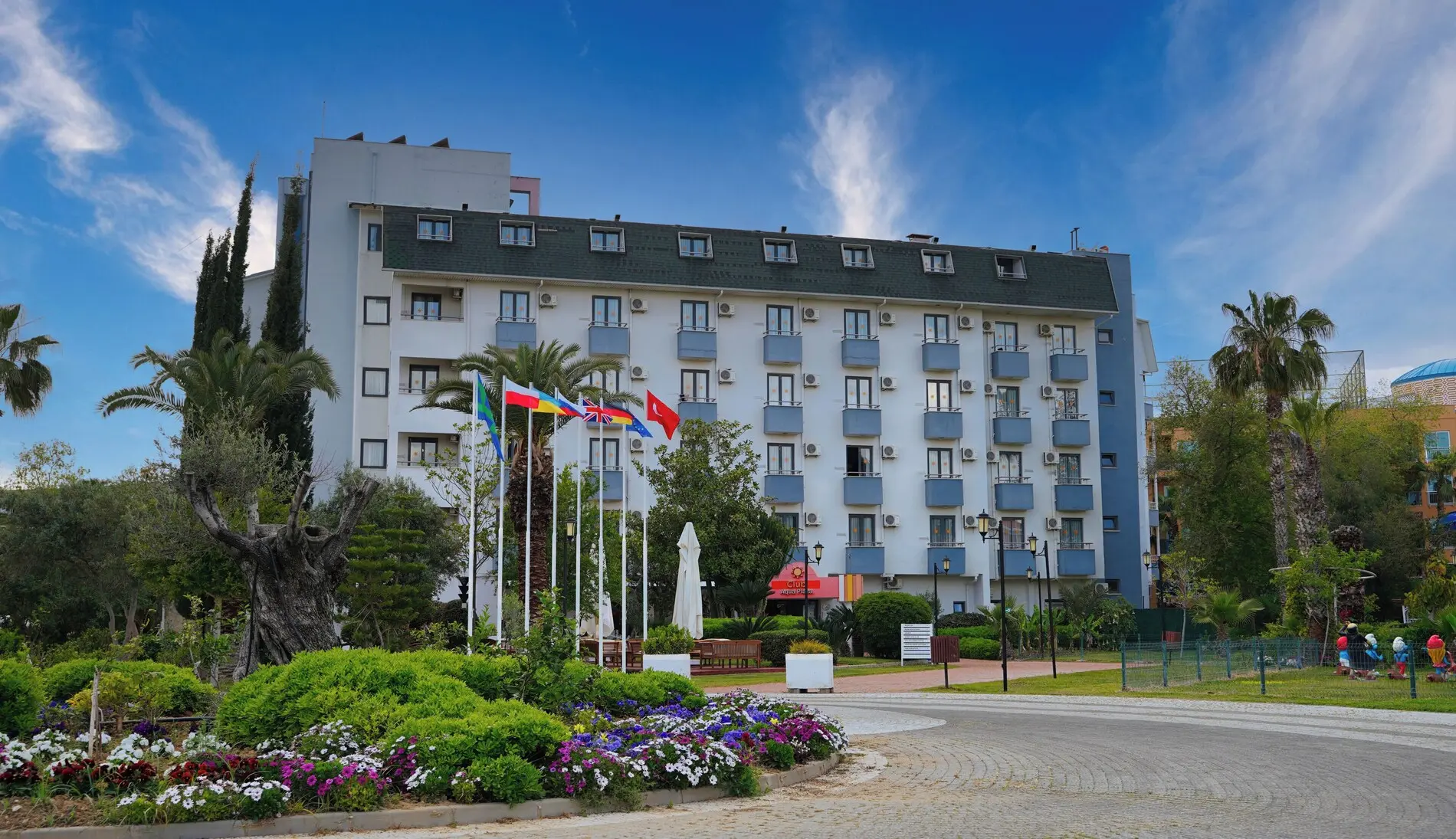 Фасадата на хотела с поддържани цветни лехи и палми пред входа – Club Aqua Plaza, Алания