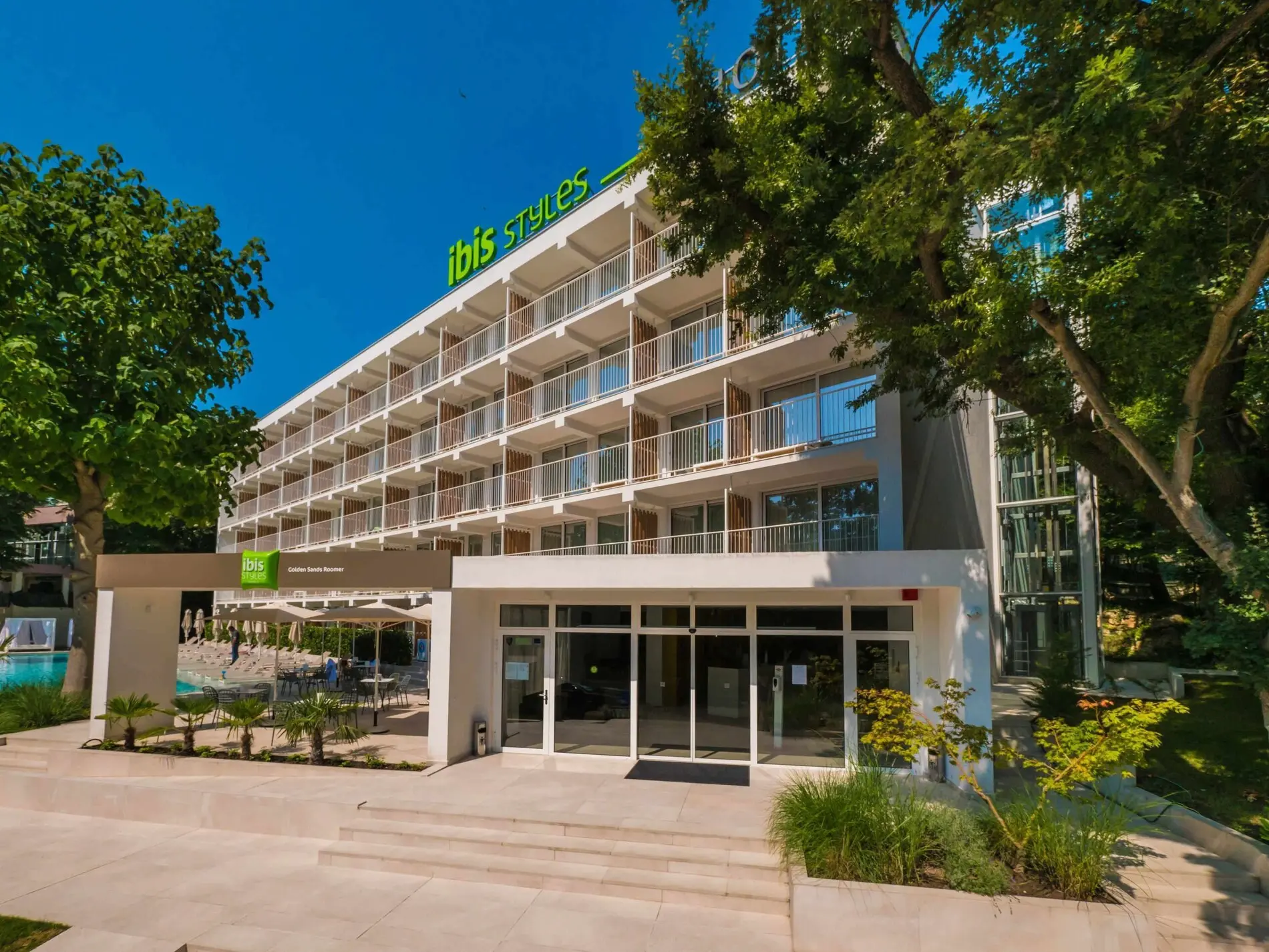 Входът и фасадата на пететажния хотел, заобиколени от зелени дървета – Ibis Styles Roomer, Златни пясъци