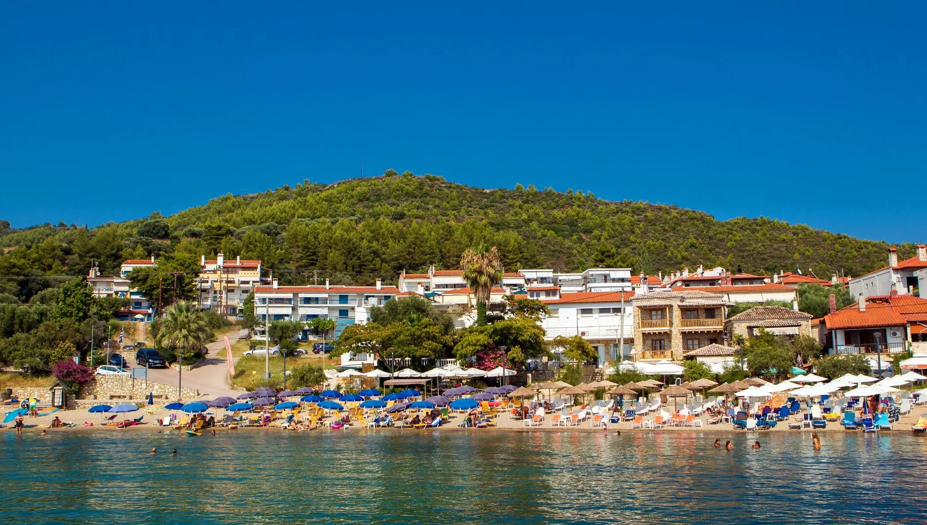 Селото и плажът с чадъри под зеления хълм – Xenios Loutra Village Beach, Касандра