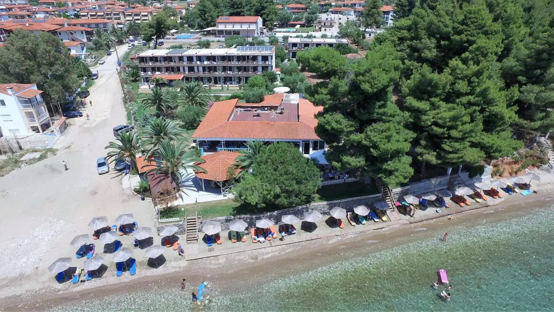 Дрон кадър на плажа с чадъри, боровете и сградите – Porto Matina, Ситония