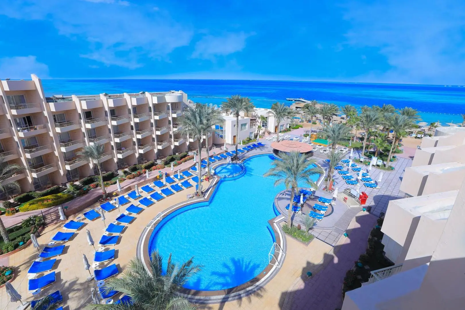 Извит басейн със сини шезлонги и палми пред сградата на хотела – Sea Star Beau Rivage Hotel, Хургада