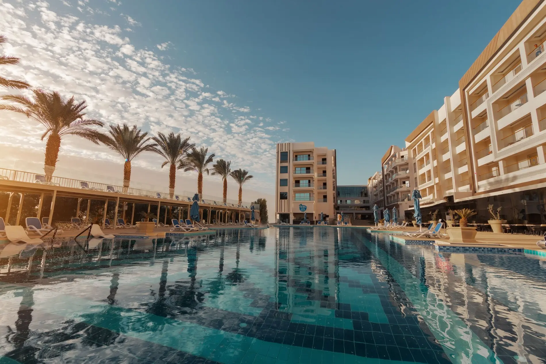 Дълъг басейн с тюркоазена вода, палми и сградите на комплекса – Bellagio Beach Resort & Spa, Хургада