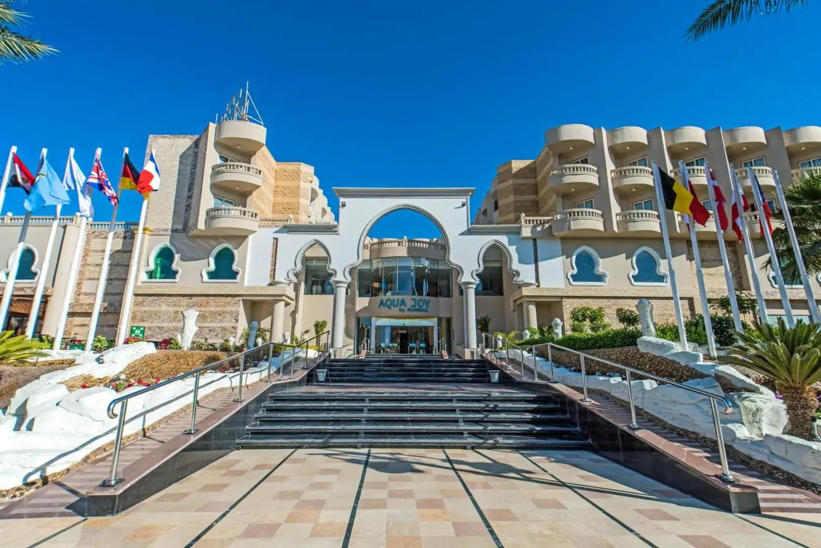 Входно стълбище и бяла фасада с арки и знамена пред главната сграда – Sunrise Aqua Joy Resort, Хургада