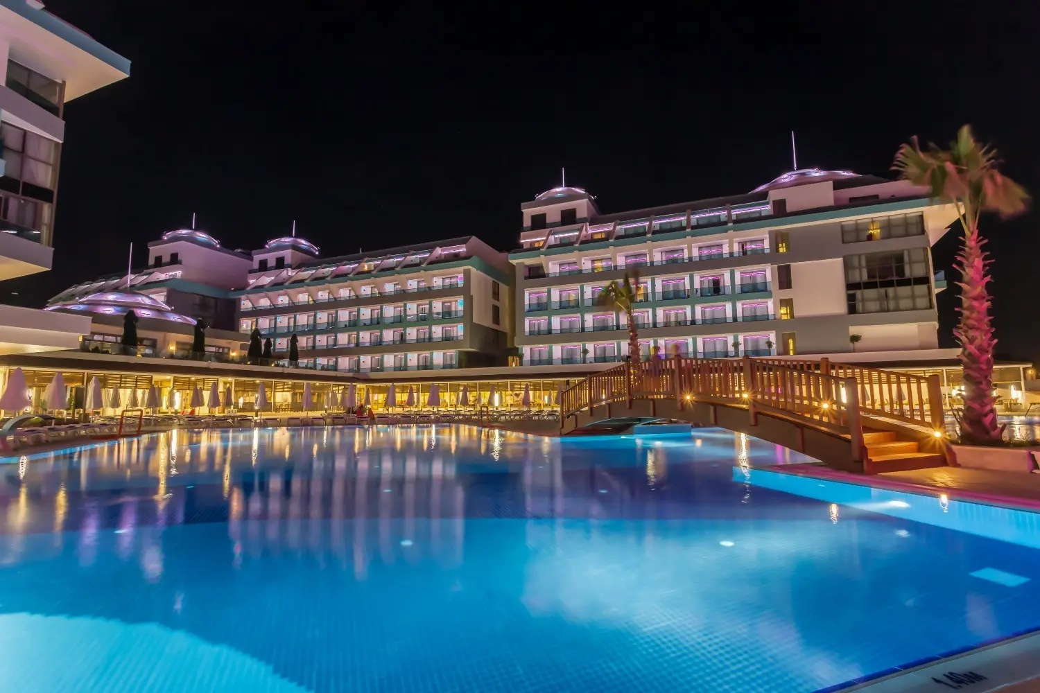 Осветени сгради на хотела зад басейна с дървен мост – Sensitive Premium Resort & Spa, Белек