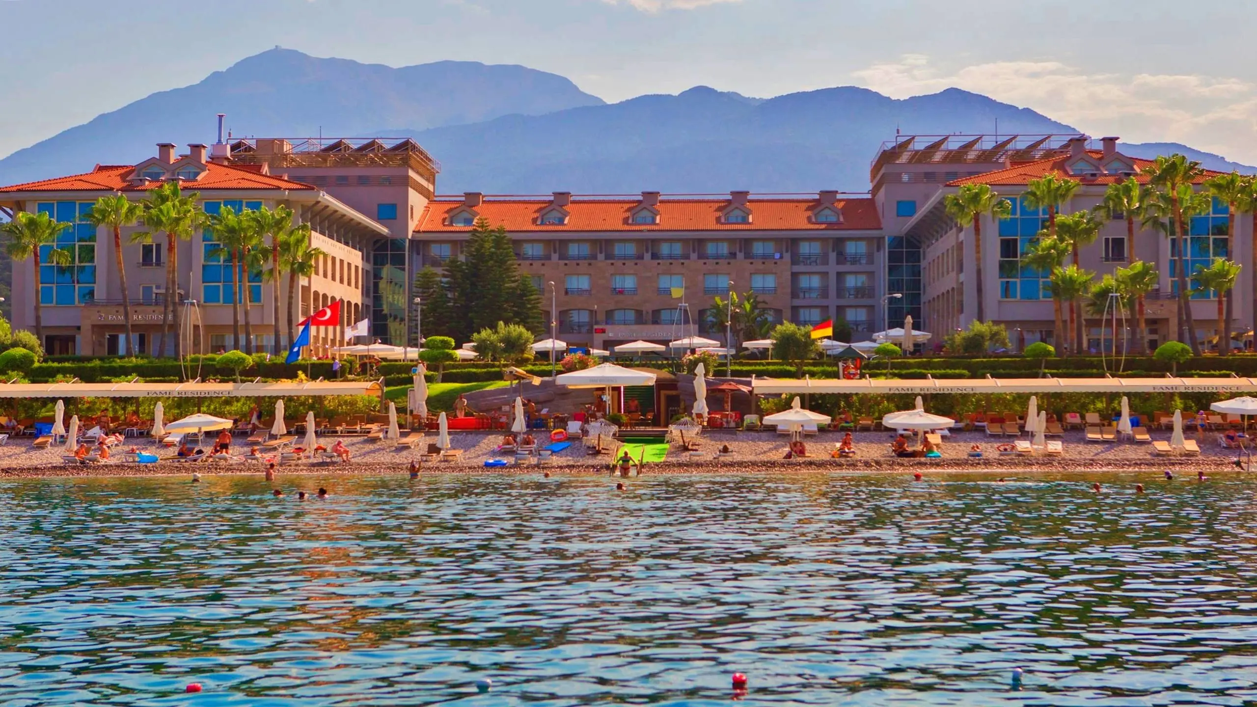 Хотелът с плажа и планините отвъд, гледан от морето – Fame Residence Kemer & Spa, Кемер