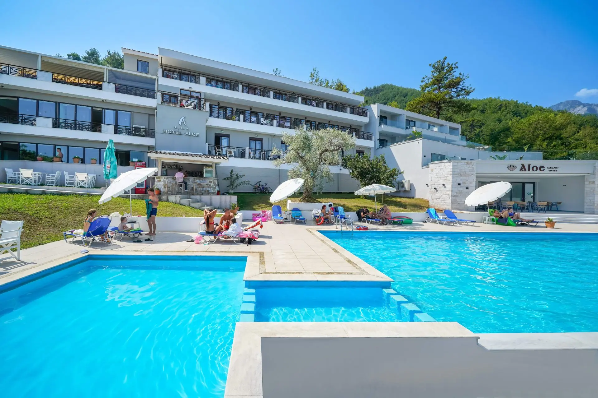Басейн с гости и чадъри пред бялата сграда на хотела – Aloe Hotel, Тасос