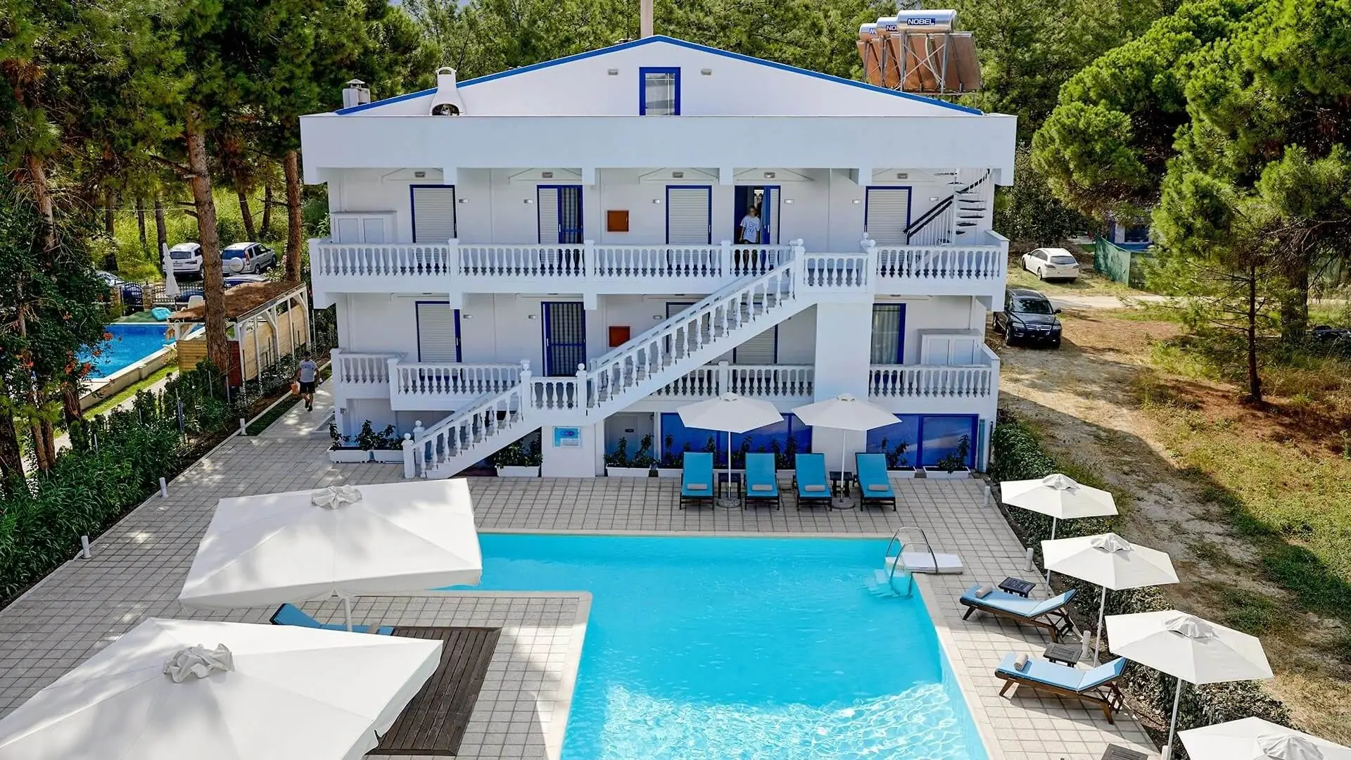 Бялата сграда със сини балкони и басейнът пред нея, снимани отвисоко – Kazaviti Hotel, Тасос