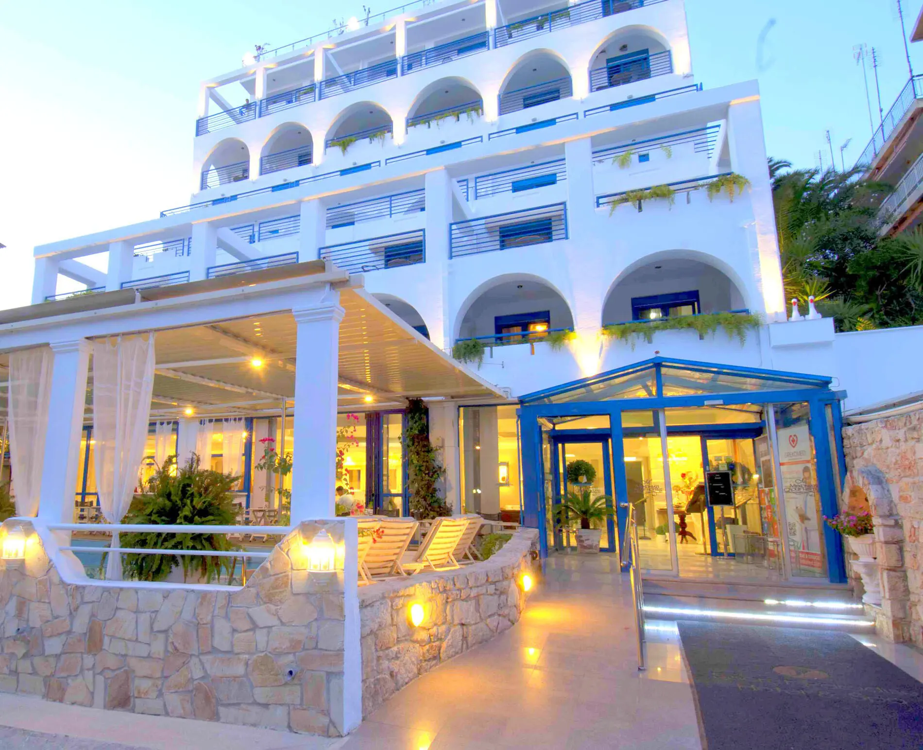 Осветена бяла фасада с арки и стъклен вход вечер – Secret Paradise Hotel & Spa, Неа Каликратия